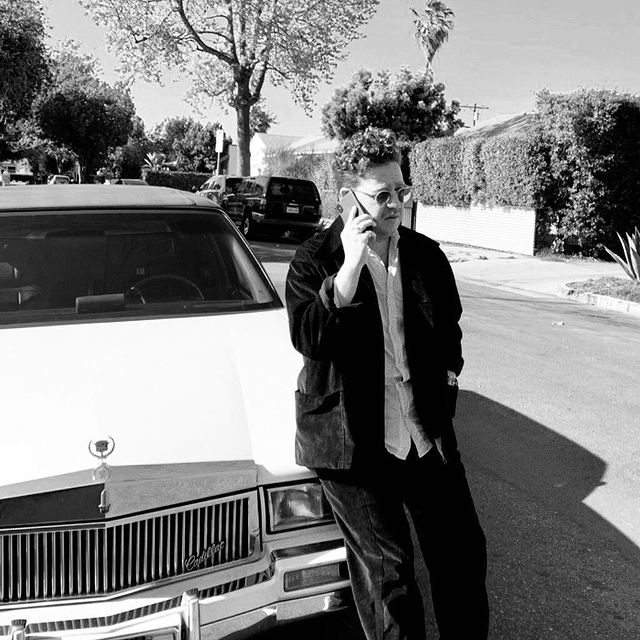 Trevor Einhorn in black pants and jacket talking in phone in front of his luxury car.